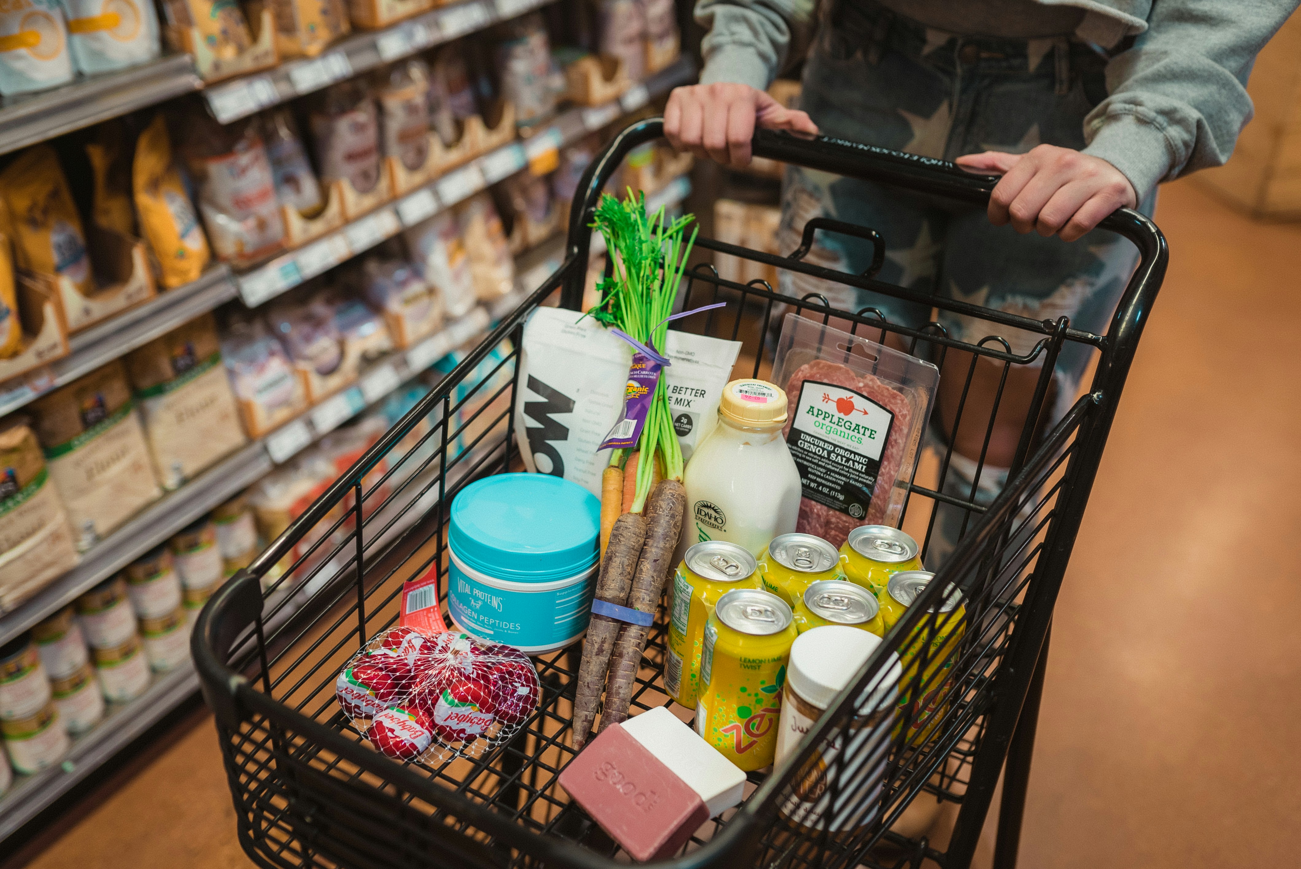 Shopping cart of groceries, including carrots, cans, and Applegate salami, in a store aisle
